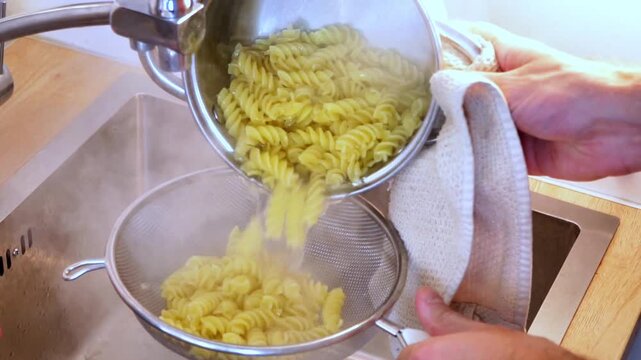 Close up hot cooked pasta pouring from pot into metal colander draining boiling water steam rising home kitchen cooking process