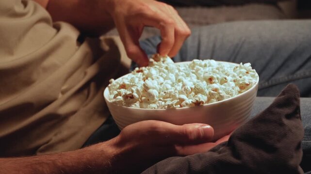 Close-up man holding bowl of fresh popcorn on lap cozy home movie night relaxed film watching evening leisure comfort snack