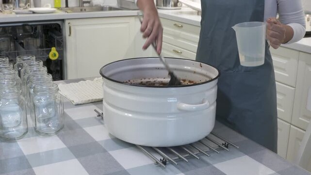 A woman in an apron stirs ingredients in a large white pot while holding a measuring jug. She prepares homemade preserves on a table lined with empty glass jars in a modern kitchen.