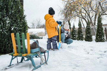 Mother and child playing in a snowy backyard, building a snowman with sled and shovel; cozy winter...