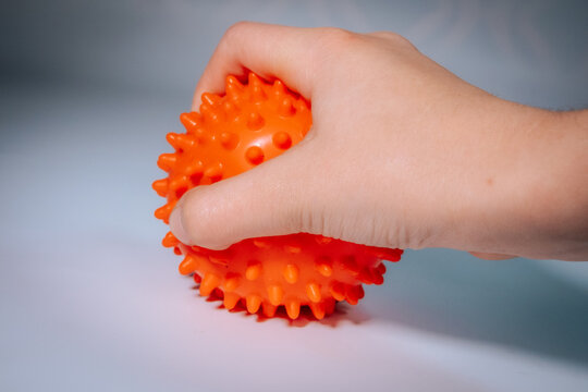 A close-up of a hand firmly gripping a textured orange, spiky massage ball on a smooth surface, seen with soft lighting.