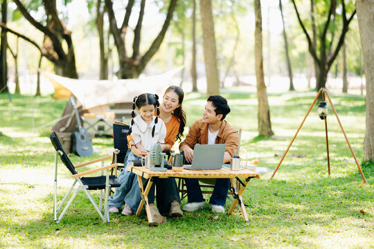 Joyful Asian family camping in a sunny garden, bonding over BBQ, tech, and laughter. Parents and daughter enjoy outdoor - Powered by Adobe