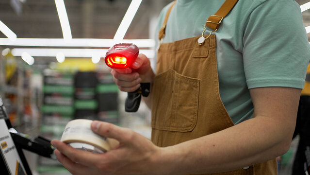 Construction worker scanning packaging tape barcode, swiping credit card at self-service hardware store checkout, busy retail environment capturing retail technology interaction