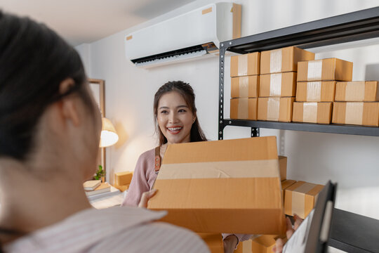Two young women are working together in a small business office, surrounded by shelves of packages and documents. They discuss tasks, check inventory, and prepare parcels with teamwork and focus.
