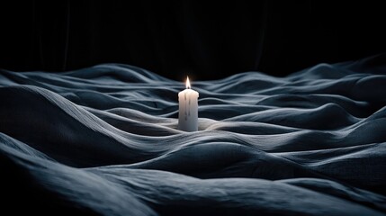 Naklejka premium A solitary white candle burns with a bright flame, casting a warm glow on a dark, textured surface that resembles rolling waves or dunes. The scene is dimly lit