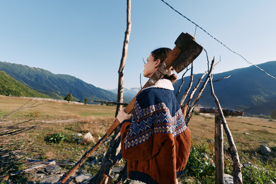 Woman with axe standing in rural countryside field near a wooden fence, wearing patterned sweater and carrying wood for chopping in mountains valley, outdoor rustic lifestyle portrait. - Powered by Adobe