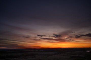 Vibrant twilight sky displaying a rich gradient of orange, red, and purple hues above a dark, silhouetted rural landscape, creating a dramatic and serene natural scene
