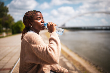Young black woman is drinking water on sunny day. She is sitting at quay by the river.