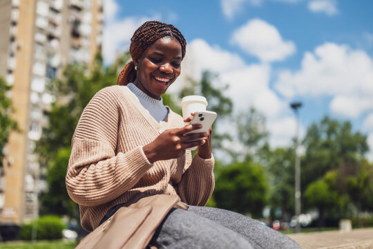 Cheerful young black woman sitting in a park, texting on her smartphone while enjoying a coffee to go on a sunny day.