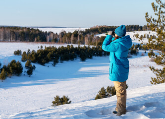 young woman in a blue jacket stands on a snowy landscape, using binoculars to observe the distant scenery of trees and fields, research in nature for ecological and conservation purposes, birdwatching
