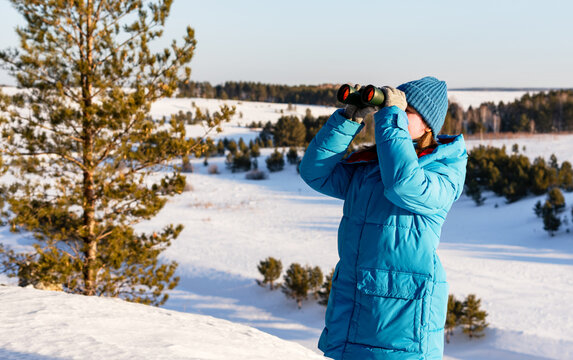 young Caucasian woman with wearing a blue winter coat and hat, uses binoculars in a snowy landscape with trees and hills in the background, Birdwatching and environmental research - Powered by Adobe