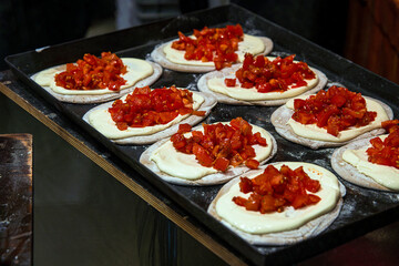 Fresh flatbreads arranged on a tray, each covered with creamy sauce and topped with chopped tomatoes or paprika mix at a holiday street food market.
