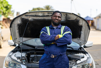 Proud African mechanic, successful small business owner in blue overalls stands confidently in front of a white car with its hood open at his workshop, SME © Roger