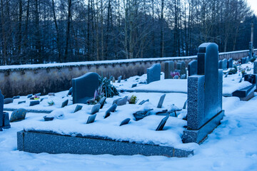 Snowy cemetery scene with old stone wall, cross, and metal gate, set against backdrop bare trees...
