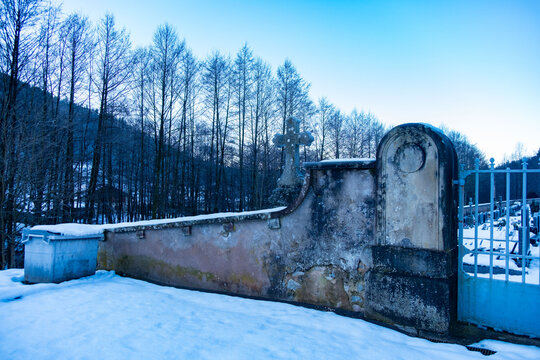 Snowy cemetery scene with old stone wall, cross, and metal gate, set against backdrop bare trees and cold blue sky, evoking peace, memory, and quiet beauty of winter