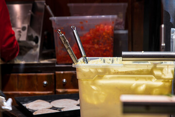 Preparation area with creamy sauce container and chopped vegetables at a festive market food stall. Tools, ingredients, and flatbreads ready to be topped,
