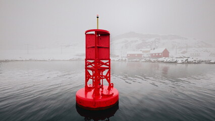 A vibrant red buoy floats in calm waters, surrounded by a foggy atmosphere. Snow covered landscapes and distant structures add an aura of tranquility to the coastal view. © icetray