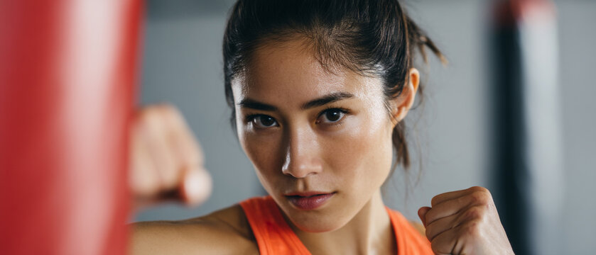 Confident Asian female athlete in orange sportswear practicing boxing in gym, focused expression, close up portrait, fitness training, determination, strength, motivation