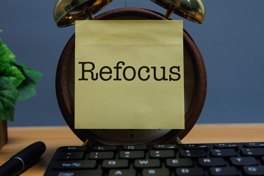 A yellow sticky note with the word &ldquo;Refocus&rdquo; on a clock face at a work desk, symbolizing a simple wellness concept of mindful breaks and regaining concentration.