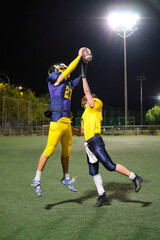 American football players in uniform jumping for a catch at night under stadium lights