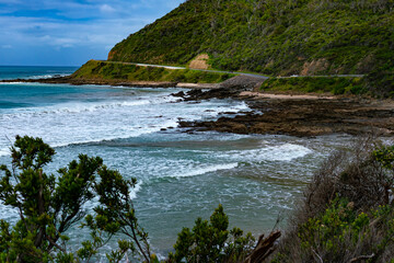View of surf crashing against the rocks on the beach with the Great Ocean Road and the green mountain, Victoria, Australia.