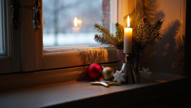 Cozy winter window sill with a lit candle pine branches and festive christmas ornaments casting warm light on a snowy day First Sunday of Advent