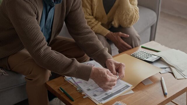 Tilt down shot of senior Caucasian couple managing finances and going through bills with calculator on sofa, showing concern over household expenses in warm living room