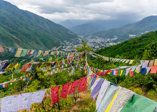 Aerial view of fluttering prayer flags leading the eye towards the distant town nestled between verdant hills under a cloudy sky, Thimphu, Thimphu, Bhutan.