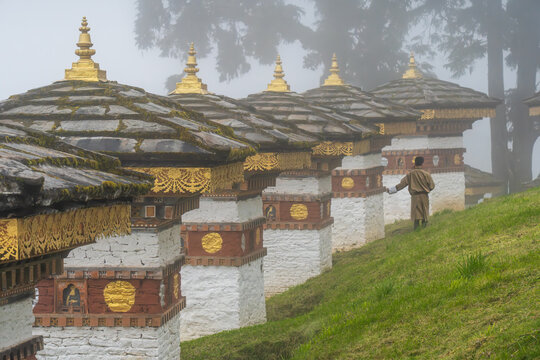 View of the 108 memorial chortens stand in orderly rows, a man walking past them on a misty day, their golden spires piercing the fog, Dochula, Thimphu, Bhutan.
