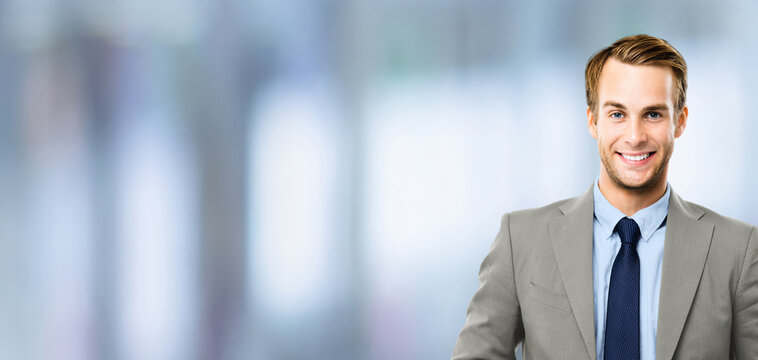 Portrait - happy smile confident business man in grey suit jacket, blue shirt, tie, against blurred office background. Businessman, bank manager, lawyer, teacher. Banner image with free ad text space