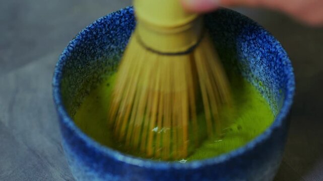 A close-up shot of a bamboo whisk stirring vibrant green matcha in a textured blue bowl. The motion creates a smooth, frothy blend, highlighting the traditional Japanese tea preparation process.