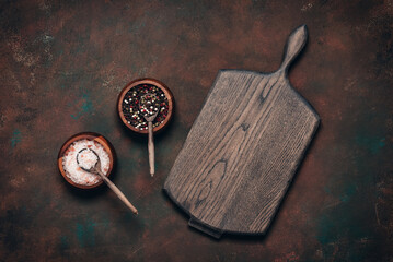 Culinary dark background with empty wooden cutting board and spices, pink salt, peppercorns, top view, flat lay