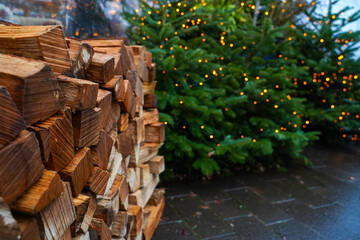 Stacked firewood with lit Christmas trees in outdoor winter setting, glowing string lights in background. Natural seasonal atmosphere, rustic holiday setting, Christmas, New Year