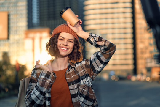 Woman coffee smile urban shopping beanie candid: young woman holding takeaway cup above head at golden hour, warm sunlight, authenticity, mindful living.
