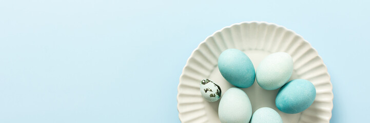 A collection of decorated blue Easter eggs sits on a white plate, showcasing various shades and patterns. The light background enhances the festive atmosphere of Easter, banner