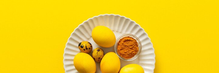 Bright yellow Easter eggs arranged on a white plate with quail eggs and a small bowl of turmeric add a vibrant touch to the holiday decorations and festivities, a way to dye eggs yellow, banner