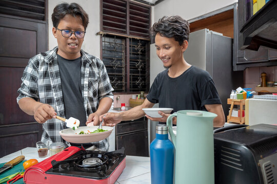 Two Indonesian southeast asian men are cooking together in a kitchen. Cooking in a frying pan on a gas stove, while holding a plate tasting the dish with a spoon. Cooking activity - Powered by Adobe