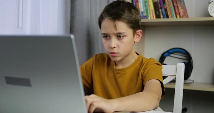 Child boy plays computer game on laptop at home. Child focuses on screen and reacts to gameplay. Young kid gamer enjoys digital entertainment. Modern technology creates immersive gaming atmosphere.