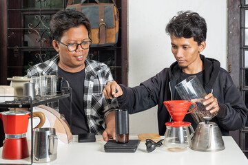 Two Indonesian southeast asian men are preparing coffee. Pouring coffee beans into a manual coffee...