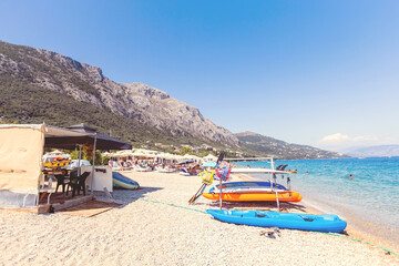 A lively pebble beach in Corfu, Greece, bustling with sunbathers, kayaks, and umbrellas against a backdrop of rugged mountains and turquoise sea.