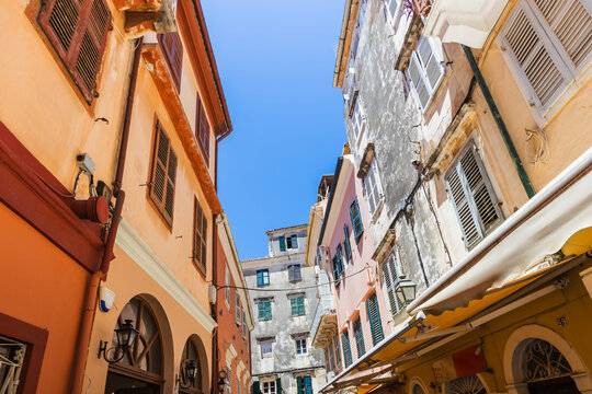 Sun-drenched narrow street in Corfu, Greece, framed by pastel buildings, wooden shutters, and clear blue sky.