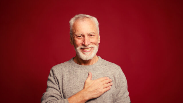 Happy grateful elderly bearded man holding hand on chest looking at camera on red background