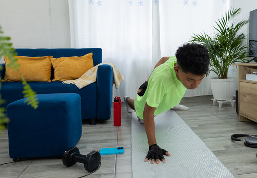 Dark skinned teenager doing a one handed push up in his living room, supported on an exercise mat. Strength exercise and home workout focused on control and endurance
