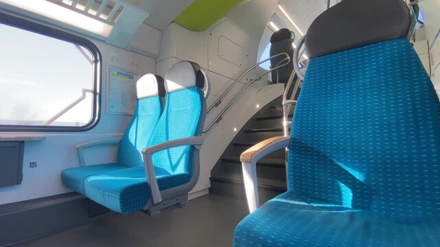 A modern, bright interior view of a double-decker European train, showcasing blue seats, a staircase to the upper deck, and large windows. The scene represents travel and transportation.