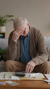 Vertical medium slowmo of distressed Caucasian senior man in glasses reviewing bills and counting expenses on sofa, managing personal finances and budgeting with concern and focus