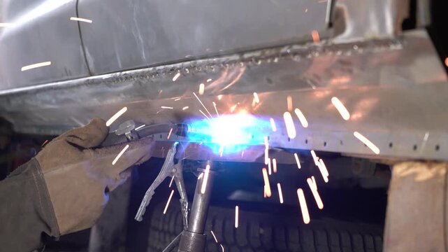 A technician performs metal welding on a car body in an auto repair shop. The process involves joining two metal pieces together and creating sparks when a torch heats the surface.