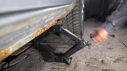 A person lifts a car using a jack to change a tire. Tools and parts are scattered around, highlighting the repair work taking place. The workshop is dimly lit, focusing on the task at hand.
