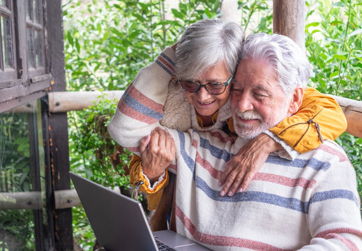 Cheerful senior couple using laptop together sitting on a rustic outdoor terrace. Dressed in winter sweaters hugging in natural mountain setting