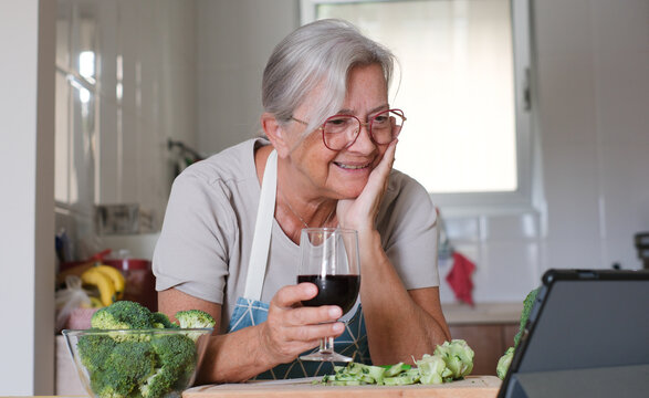 Elderly woman wearing apron using digital tablet while enjoying a red wineglass after chops fresh broccoli on a kitchen counter. Concept of healthy meal preparation, fresh vegetables, home cooking