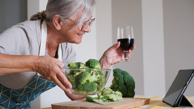 Elderly woman wearing apron using digital tablet while enjoying a red wineglass after chops fresh broccoli on a kitchen counter. Concept of healthy meal preparation, fresh vegetables, home cooking